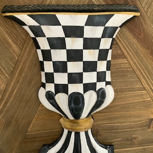 May include: Black and white checkered ceramic vase with a gold trim and a black rope detail around the top. The vase has a classic urn shape with a fluted base.