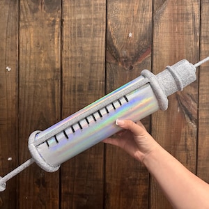 May include: A large, decorative syringe prop with a silver glitter finish. The body of the syringe has a holographic, iridescent sheen. The plunger and needle are also silver. The syringe is held against a wooden background.
