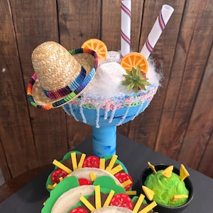 May include: A blue margarita glass with a straw, orange slices, and a small sombrero on top. The glass is filled with white cotton candy and surrounded by three tacos and a small bowl of guacamole.