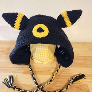 Pode incluir: Chapéu de crochet preto com orelhas amarelas e um círculo amarelo no topo. O chapéu tem um cordão trançado longo preto e amarelo com borlas.