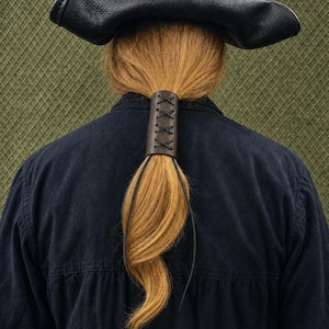 Puede incluir: Un accesorio para el cabello de cuero marrón oscuro con un diseño de cordones cruzados. El elemento cilíndrico está asegurado alrededor de una coleta de cabello castaño claro. Un sombrero de tres picos de cuero negro descansa sobre la cabeza.