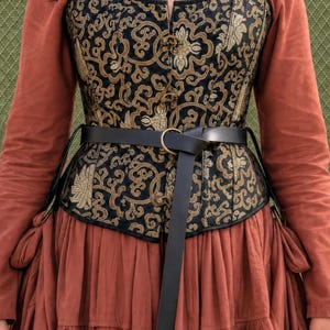 May include: A structured corset featuring a black and gold floral pattern with a front closure, worn over a long-sleeved terracotta-colored garment. A thin black leather belt with a circular metal buckle is cinched around the waist of the corset.