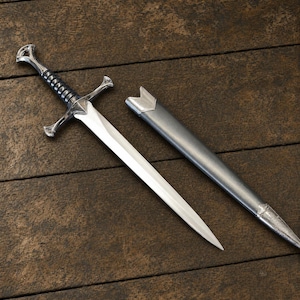 May include: A silver-toned metal sword with a pointed blade, a crossguard, and a black ribbed grip, accompanied by a matching metallic gray scabbard. The sword features a pommel at the end of the handle and a tapered blade design.