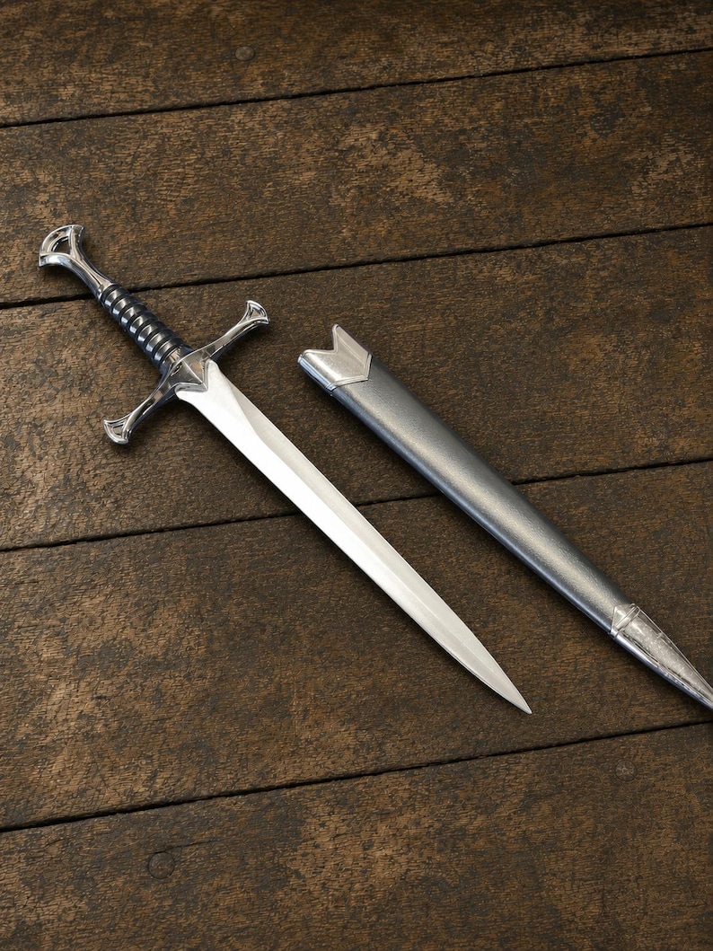 May include: A silver-toned metal sword with a pointed blade, a crossguard, and a black ribbed grip, accompanied by a matching metallic gray scabbard. The sword features a pommel at the end of the handle and a tapered blade design.
