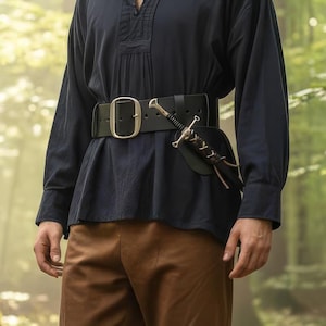 May include: A black leather belt with a large silver buckle. A small sword in a black sheath is attached to the belt. The person is wearing a dark blue long-sleeve shirt and brown pants.