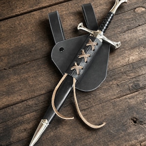 May include: A silver-toned sword with a black grip and crossguard, housed in a black leather scabbard. The scabbard features a decorative tan leather lace-up detail and is attached to a black leather belt frog with a single metal rivet.