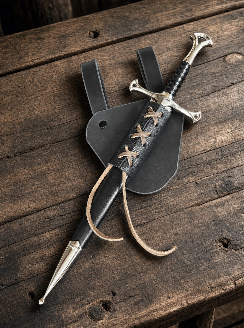 May include: A silver-toned sword with a black grip and crossguard, housed in a black leather scabbard. The scabbard features a decorative tan leather lace-up detail and is attached to a black leather belt frog with a single metal rivet.