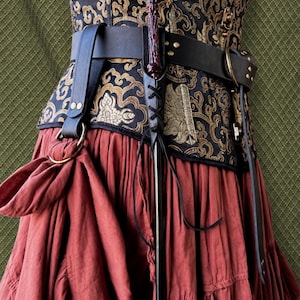 May include: A black leather belt with a brass ring and a matching leather holster holding a dark red, textured wand. The belt is worn over a black and gold patterned corset and a long, solid rust-colored skirt with gathered fabric details.