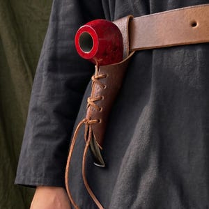 Puede incluir: Una funda de cuero marrón oscuro para pipa con diseño de cordones, sujeta a un cinturón de cuero ancho a juego. Una pipa de tabaco roja brillante con boquilla negra está asegurada dentro de la funda, sobre una prenda negra de manga larga.