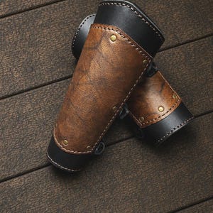 May include: A pair of arm bracers crafted from black and brown leather. The bracers feature a textured brown leather center panel secured to a smooth black leather base with small brass-colored metal rivets and visible stitching along the edges.