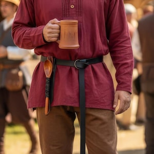 Puede incluir: Una túnica burdeos de manga larga, un cinturón negro y pantalones marrones. Una cuchara de madera está metida en una funda negra sujeta al cinturón. Una jarra de cerámica marrón se sostiene en la mano.