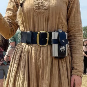 May include: A tan-colored dress with long sleeves and a black belt with a gold buckle. A black case with silver accents is attached to the belt. The dress has a pleated skirt and detailed embroidery on the bodice.