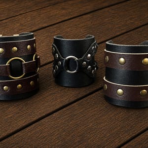 May include: Three wide leather cuff bracelets. The left cuff is dark brown with brass studs and two central rings. The centre cuff is black with silver studs and a central ring. The right cuff is black with two dark brown bands and brass studs.