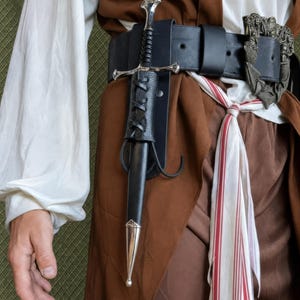 May include: A black leather dagger sheath with silver metal accents and a matching silver-hilted dagger, secured to a wide black leather belt with an ornate silver buckle. The belt is worn over a brown vest, a white shirt, and a red-striped sash.