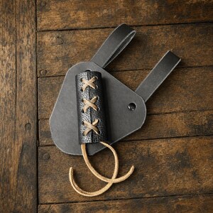 Puede incluir: Un accesorio de cintur&oacute;n de cuero negro con un soporte cil&iacute;ndrico de superficie texturizada. El soporte est&aacute; asegurado con cordones de cuero color canela en un patr&oacute;n entrecruzado e incluye dos largos lazos de cuero colgando del borde.