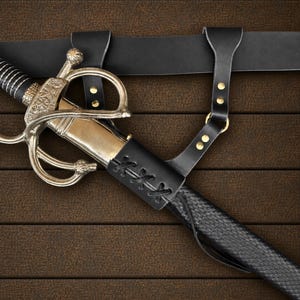 May include: A sword with a metallic gold-toned scabbard and an ornate silver-colored hilt featuring a cross-guard. The sword is secured in a black leather frog with decorative lacing and brass rivets, attached to a matching black leather belt.
