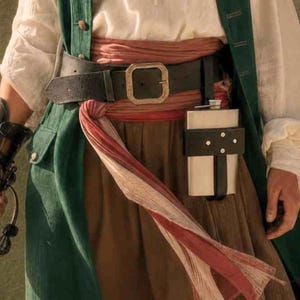 Puede incluir: Un conjunto de disfraz con un chaleco largo verde sobre una camisa blanca, una falda marrón y una faja roja ancha. Un cinturón de cuero negro con hebilla de latón se lleva sobre la faja, sujetando una petaca de metal en un soporte negro.