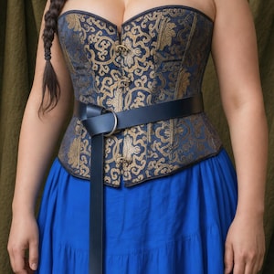 May include: A strapless corset featuring a dark blue and gold damask pattern with a front metal clasp closure. A dark blue leather belt with a silver-toned metal ring buckle is worn over the waist of the corset, paired with a bright blue skirt.