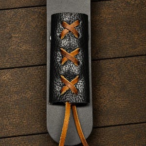 May include: A black leather sheath featuring a textured black leather overlay secured with brown leather lacing in a crisscross pattern. The lacing extends from the bottom of the overlay in two long, loose brown leather cords.