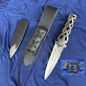 May include: A silver dagger with an elaborate handle and a serrated blade, presented with a black leather sheath and belt loop. The handle showcases an intricate Celtic knot design. A small inset shows additional daggers with coloured accents.