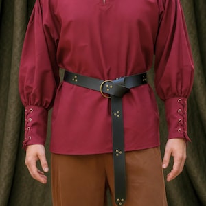 May include: A deep red long-sleeved shirt featuring a lace-up neckline with metal grommets, gathered sleeves, and cuffs with lace-up detailing. A black leather belt with a brass ring buckle and decorative brass studs is worn over the shirt.