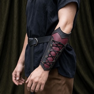 May include: A forearm bracer crafted from deep burgundy leather with black leather trim and edges. The piece features a central lacing system with black cord threaded through brass-toned metal eyelets, secured around the arm with a structured design.