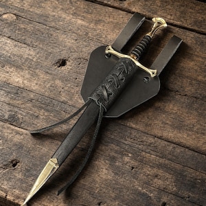 May include: A dagger with a gold-toned metal crossguard and pommel, featuring a black ribbed grip. The blade is housed in a black leather scabbard with a lace-up detail and a gold-toned metal tip, attached to a black leather belt frog mount.