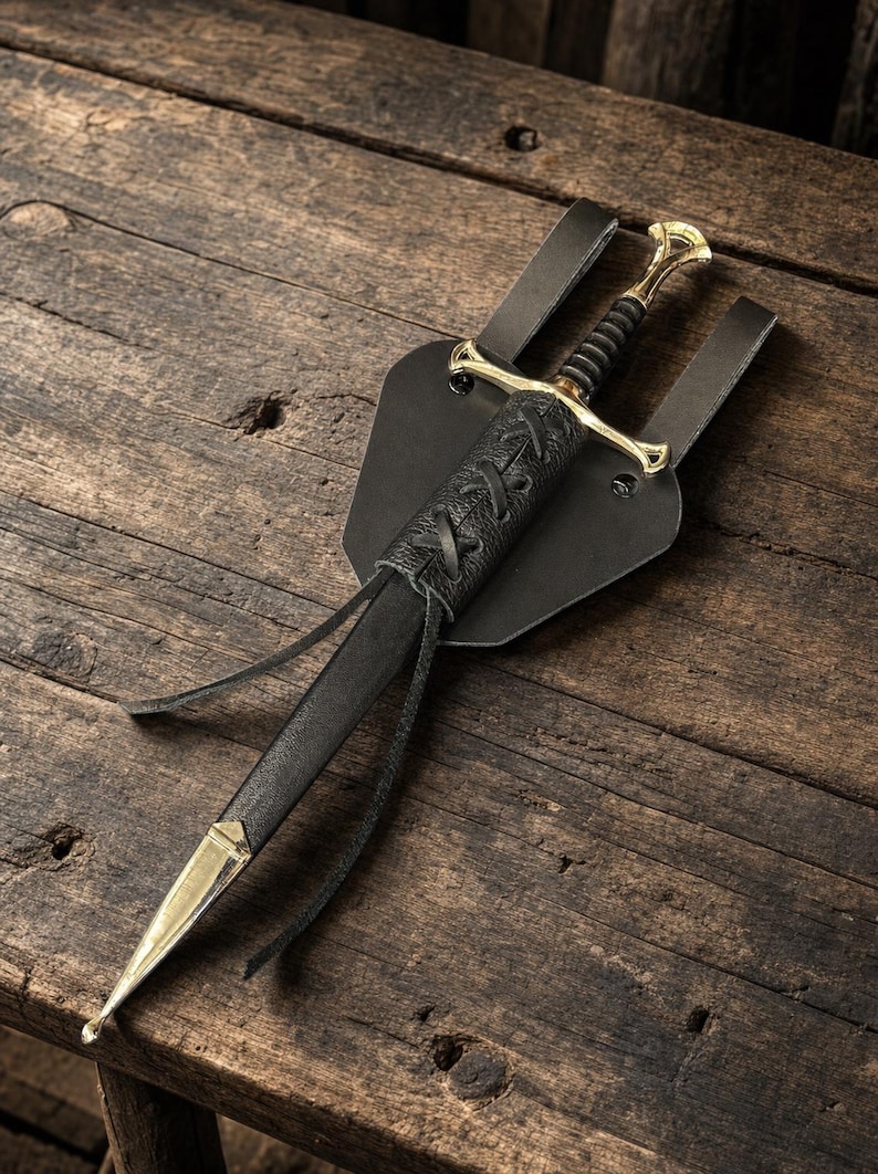May include: A dagger with a gold-toned metal crossguard and pommel, featuring a black ribbed grip. The blade is housed in a black leather scabbard with a lace-up detail and a gold-toned metal tip, attached to a black leather belt frog mount.