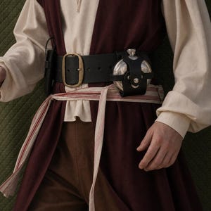 May include: A black leather belt with a brass-toned buckle, featuring a circular metal flask secured in a black leather holster. The belt is worn over a cream-colored long-sleeved shirt, a dark maroon vest, brown trousers, and a striped fabric sash.