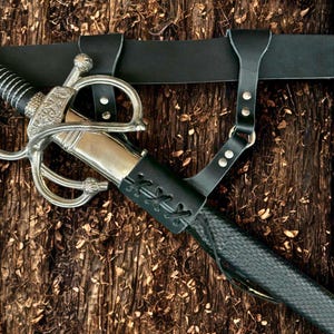 Puede incluir: Una espada decorativa con una empuñadura plateada y una vaina de cuero negro. La espada está sujeta a un cinturón de cuero negro con una correa. El fondo es una superficie de madera marrón oscura texturizada.