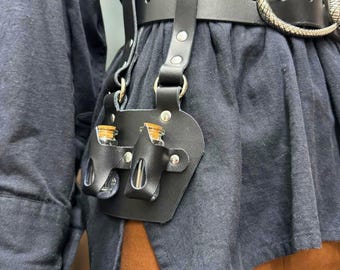 May include: A black leather belt featuring a large oval buckle with a textured metallic finish. Attached to the belt is a leather holster component holding two small clear glass vials with cork stoppers, secured by silver-toned metal rivets.
