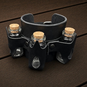 May include: A black leather cuff bracelet featuring three integrated vertical slots, each holding a small clear glass vial with a cork stopper. The leather band is secured with a metal buckle and features dark metal rivets at the vial attachment points.