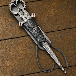 May include: A pair of silver-toned metal scissors with intricate embossed patterns and small red accents, housed in a black textured leather sheath featuring a crisscross lace-up design and long leather ties extending from the base.