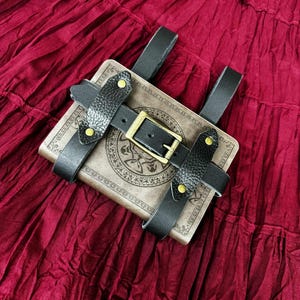 Pode incluir: Um livro castanho com um design decorativo &eacute; preso por tiras de couro preto e uma fivela. As tiras s&atilde;o presas com rebites dourados. O livro e as tiras s&atilde;o colocados sobre um fundo de tecido vermelho escuro.