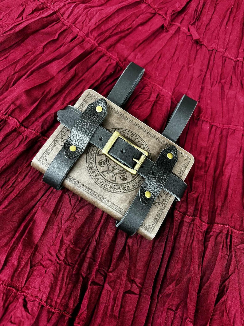 Pode incluir: Um livro castanho com um design decorativo &eacute; preso por tiras de couro preto e uma fivela. As tiras s&atilde;o presas com rebites dourados. O livro e as tiras s&atilde;o colocados sobre um fundo de tecido vermelho escuro.