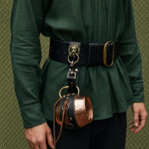 May include: A wide black leather belt featuring a brass lion head buckle attachment and a matching brass oval buckle. A copper cup with a hammered texture is suspended from the belt by a black leather strap and a brass ring mechanism.