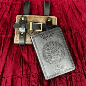 May include: A brown and black leather book holder with a gold buckle and two books. The books feature detailed embossed designs. The holder has black straps and gold rivets. The background is a deep red fabric.