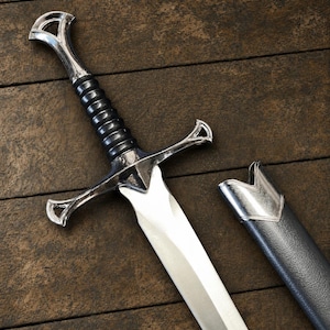 May include: A silver-toned metal sword with a black ribbed grip and a polished crossguard. The sword features a pointed blade and is accompanied by a matching black scabbard with a silver-toned metal throat piece at the top edge.