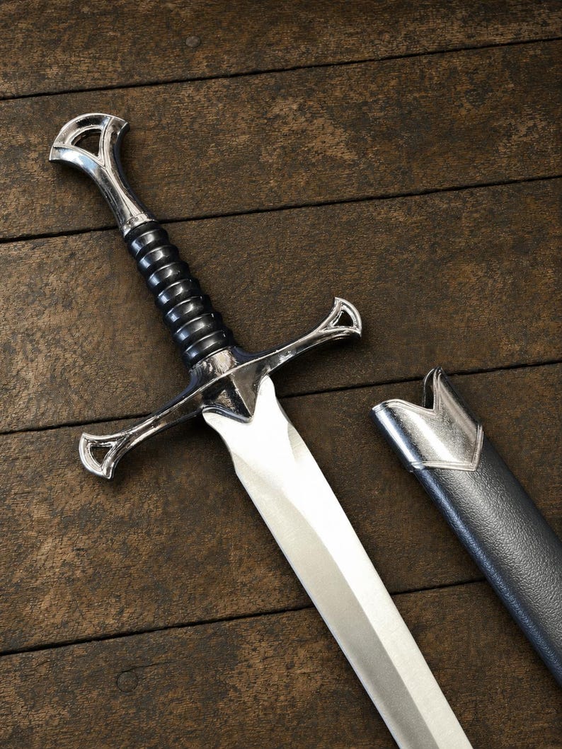 May include: A silver-toned metal sword with a black ribbed grip and a polished crossguard. The sword features a pointed blade and is accompanied by a matching black scabbard with a silver-toned metal throat piece at the top edge.
