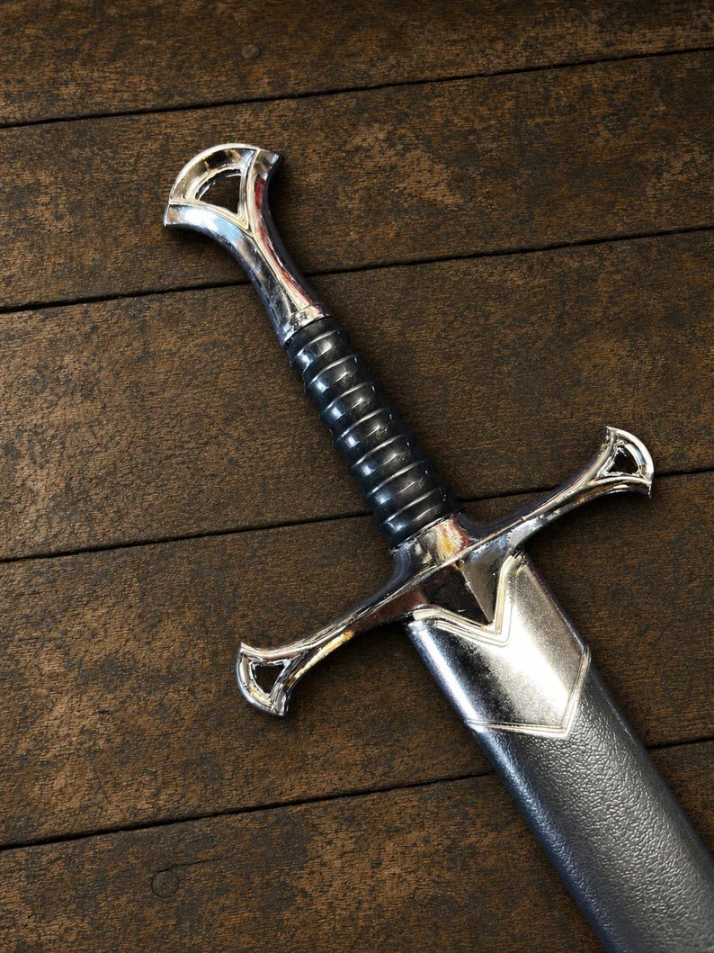 May include: A sword with a polished silver-tone metal pommel, crossguard, and throat. The handle features a black, ribbed grip. The sword is partially encased in a black, textured scabbard with a silver-tone metal fitting at the top.