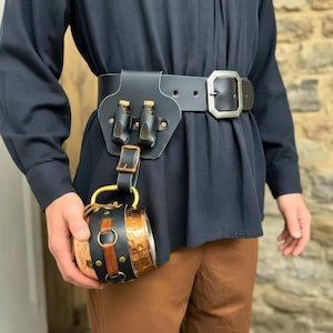 May include: A black leather belt with a silver buckle, featuring a leather pouch with two small bottles and a copper and leather barrel-shaped container. The container is held by a gold-coloured metal ring.