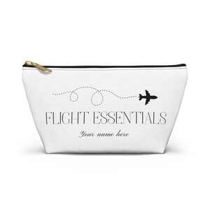 Peut inclure: Trousse de voyage blanche avec fermeture éclair dorée et texte noir "FLIGHT ESSENTIALS" avec une ligne pointillée et un avion. En dessous, le texte "Your name here".