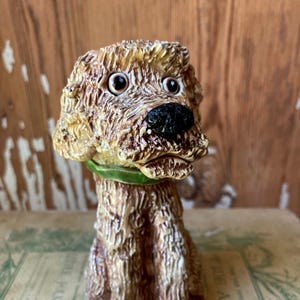 May include: A ceramic dog figurine with a textured, brown and tan coat, black nose, and large, expressive eyes. The dog wears a green collar. The figurine is sitting on a vintage book.