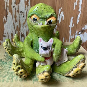 May include: A whimsical ceramic sculpture of a green creature with large eyes and sharp teeth, holding a small pink dog. The sculpture has a textured surface and is painted with yellow and orange accents. The dog has a red collar.