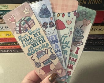 May include: Four festive bookmarks with holiday-themed designs are held in a hand. Each bookmark features a different illustration, including a stocking, a hat, mittens, and a mug. The text on the bookmarks reads "Dear Santa, Fill My Stocking With Books" and other holiday phrases.