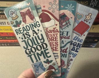 May include: Four festive bookmarks with holiday-themed designs are held in a hand. Each bookmark features a different design, including a Santa hat, snowflakes, and gift boxes. The text on the bookmarks reads "Reading is a Jolly Good Time" and other holiday phrases.