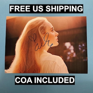 Puede incluir: Fotografía firmada de una mujer con cabello largo y castaño rojizo, mirando hacia la derecha. La foto tiene una firma en tinta azul y el texto "COA INCLUDED". El fondo está borroso con tonos cálidos.