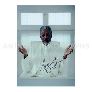 May include: A photograph of Morgan Freeman in a white suit, seated and gesturing with open hands. The image is signed with a black marker. The background is a light-colored wall with a window.