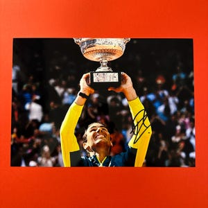 May include: A tennis player lifts a silver trophy above his head, smiling and looking up at the trophy. He is wearing a yellow and green shirt and is surrounded by a cheering crowd.