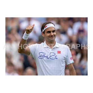 May include: A white tennis shirt with a blue signature and the UNIQLO logo. The athlete is wearing a white headband and wristband, giving a thumbs up. The background is blurred, suggesting a sporting event.